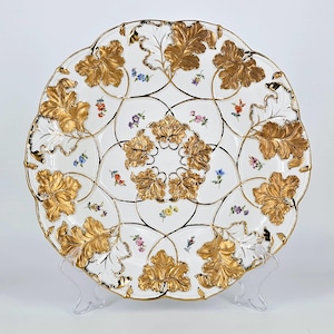 May include: A white porcelain plate with a gold leaf design and floral accents. The plate has a scalloped edge and is decorated with a repeating pattern of leaves and flowers.