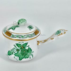 Puede incluir: Salsera de porcelana blanca con tapa y asa, adornada con motivos florales y de hojas verdes. La olla presenta un ribete dorado y un pomo verde en la tapa. El asa también está decorada con detalles dorados.