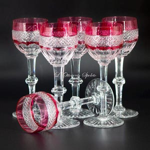Può includere: Un set di sei calici da vino in cristallo con un motivo a taglio diamante sulla coppa e una fascia rosso scuro sul bordo. Ogni calice ha uno stelo sfaccettato e una base circolare. Il testo L'Ottocento Spoleto è visibile al centro dell'immagine.