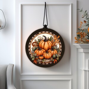 May include: A round wooden sign with a black and white checkered border. The center of the sign features a painting of orange pumpkins with fall foliage and wheat stalks. The sign is hanging from a black cord.