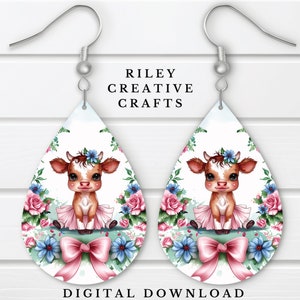 Könnte beinhalten: Ein Paar Hängeohrringe mit einer Cartoon-Kuh, die ein rosa Tutu und eine Blumenkrone trägt. Die Kuh ist von rosa und blauen Blumen und einer rosa Schleife umgeben. Die Ohrringe sind weiß und haben eine Tropfenform.