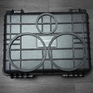 May include: A black rectangular hard-shell protective case with a grid pattern and the text Apache Rugged Utility. Three flat black circular rings of varying sizes are positioned on top of the case, with one smaller ring above two larger rings.