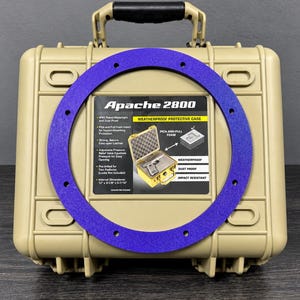 May include: A tan Apache 2800 weatherproof protective case with a black handle and a large, flat, purple circular gasket featuring multiple pre-drilled holes positioned over the front label which details internal dimensions of 12 inches by 9-1/8 inches by 5-7/16 inches.
