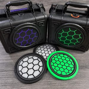 May include: Two black rectangular hard-shell cases with top handles and circular speaker grilles featuring purple and green honeycomb patterns. Two additional circular speaker grilles with white and green honeycomb designs are placed in front.