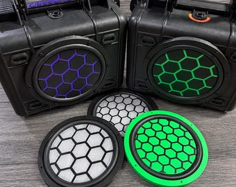 May include: Two black portable speakers with top handles and hexagonal grille patterns, one with purple and one with green accents. Two additional circular speaker grilles are placed in front, one with a white mesh and one with a bright green frame.