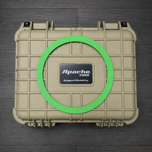 May include: A tan, rectangular, hard-shell protective case with a grid-like exterior pattern. A black rectangular label in the center displays the white text Apache 2800 Rugged Mobility. A bright green circular ring is placed on the case lid.