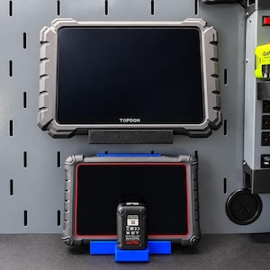 May include: A large gray Topdon diagnostic tablet mounted on a wall panel above a smaller gray Autel diagnostic tablet with a red border, which is resting on a blue plastic stand. A green GaN charger is plugged into a power strip on the right.