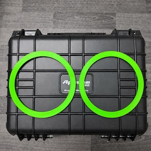 May include: Two bright lime green circular rings positioned on top of a black rectangular hard-shell protective case. The case features a grid-like molded exterior pattern and a central rectangular label with the text Apache 3800 Rugged Utility.