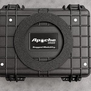 May include: A black rectangular protective hard case with a grid-patterned exterior. Centered on the lid is a circular textured ring surrounding a black rectangular label that reads Apache 1800 Rugged Mobility in white text with a white dot.