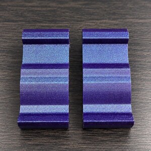 May include: Two identical, rectangular, purple-toned objects with a shimmering, glittery finish. Each piece features a series of three horizontal, rounded ridges or waves running across its surface, creating a textured, undulating profile.