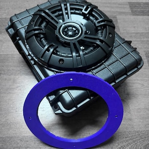 May include: A black circular audio speaker mounted within a black rectangular protective case with a textured grid pattern. In the foreground, a flat, purple circular ring with four small mounting holes is positioned in front of the speaker unit.