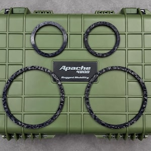 May include: An olive green rectangular hard-shell protective case with a grid pattern and a central black label reading Apache 4800 Rugged Mobility. Four black circular rings with a faceted, textured surface are arranged on the case lid.