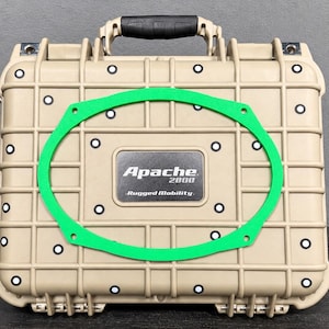 May include: A tan, rectangular, hard-shell protective case with a black handle and a grid pattern on the exterior. A bright green, oval-shaped frame is centered on the front. The case features a black label with the text Apache 2800 Rugged Mobility.