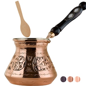 May include: A copper Turkish coffee pot with an ornate floral design and a wooden handle. The pot is sitting on a white background and a wooden spoon is floating above it.