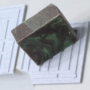 May include: A cube of handmade soap with a marbled green and brown design rests on a light grey soap dish. The soap dish has a geometric pattern. The soap has a dark brown top.