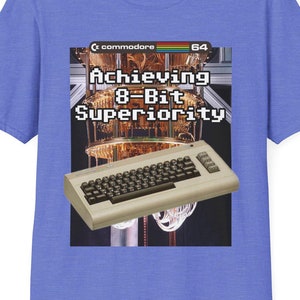 Puede incluir: Camiseta azul bígaro con un diseño gráfico de un ordenador Commodore 64 y el texto "Achieving 8-Bit Superiority". El diseño incluye un teclado de ordenador retro y un fondo estilizado.
