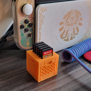May include: Orange plastic crate-style game cartridge holder with five Nintendo Switch game cartridges inside. The holder is sitting on a wooden surface next to a gold Nintendo Switch console.