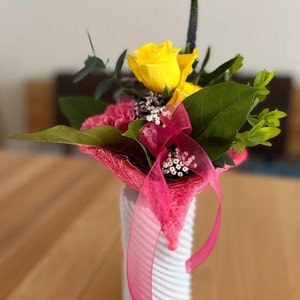 Op de afbeelding: Een klein boeket met gele rozen en roze anjers in een witte vaas met een roze lint.