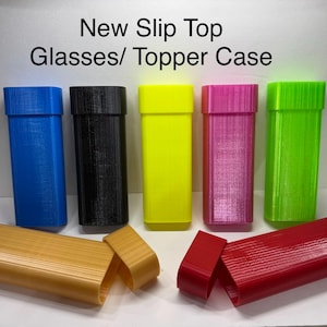 May include: A collection of colorful slip-top cases in blue, black, yellow, pink, and green. The cases are rectangular with a textured surface. The image includes the text "New Slip Top Glasses/ Topper Case".