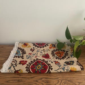 May include: Folded beige fabric featuring a floral pattern in red, yellow, blue, and orange. One edge is trimmed with white lace. The floral design includes stylized flowers and leaves.