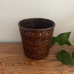 May include: A dark brown woven basket with a cylindrical shape and a tapered base. The basket features a complex pattern of horizontal bands and intricate herringbone-style weaving, finished with a smooth, rounded rim at the top opening.