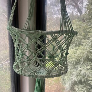 May include: A light green macrame plant hanger with a basket-like design. The plant holder is suspended by a woven cord and features a diamond-shaped pattern. Small green beads are incorporated into the design, and a tassel hangs from the bottom.