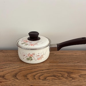 May include: A white enamel saucepan with a brown handle and floral design. The saucepan has a brown knob on the lid.
