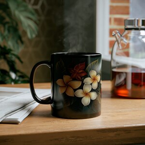 May include: A black ceramic mug with a floral design featuring white plumeria flowers and green leaves. The mug is filled with a steaming beverage.