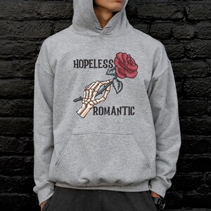 May include: A gray hooded sweatshirt with the text "HOPELESS ROMANTIC" printed on the front. A skeleton hand is holding a red rose.