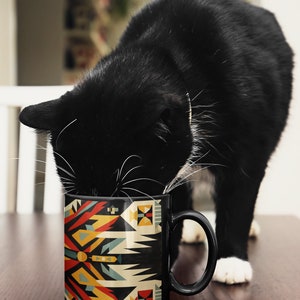 May include: A black and white cat drinking from a black ceramic mug with a colorful geometric pattern. The mug has a handle and is on a wooden surface.