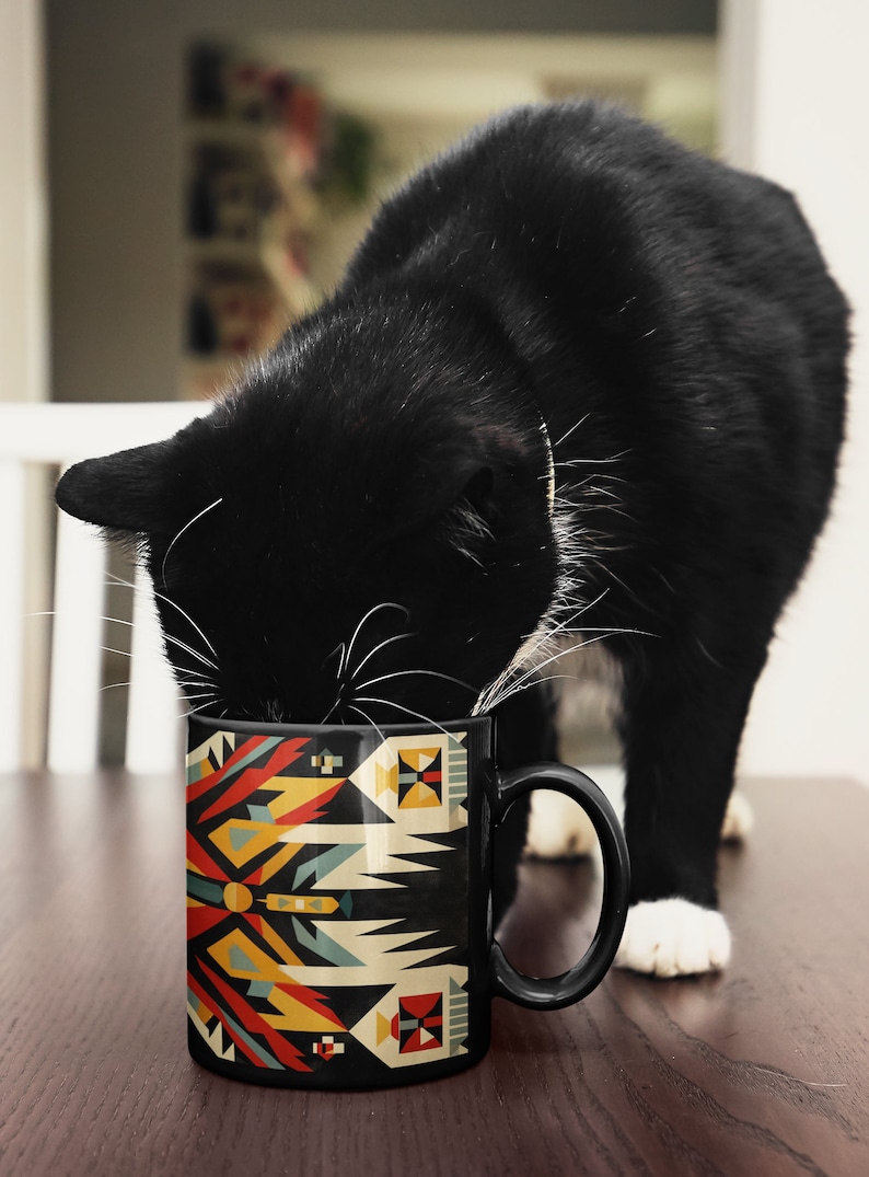 May include: A black and white cat drinking from a black ceramic mug with a colorful geometric pattern. The mug has a handle and is on a wooden surface.