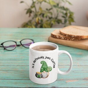 May include: White ceramic coffee mug with a green, white, and gold gnome illustration and the text "It's definitely just coffee".