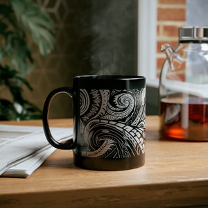 May include: A black ceramic mug with a white Polynesian-inspired design. The mug is filled with a hot beverage, and steam is rising from the top.