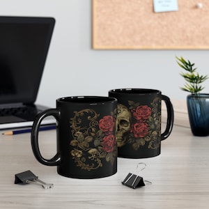 May include: Two black ceramic mugs with a skull and roses design. The mugs have a black background with gold accents and red roses. The skull is in the center of the design and is surrounded by roses.