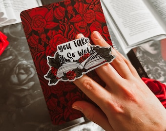 May include: A red and black journal with a rose pattern is held by a hand. A sticker with the text "you take it so well" is on the journal. Other books and a red rose are in the background. The journal has a floral design.