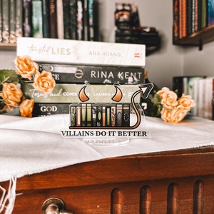 Puede incluir: Un separador de libros de acr&iacute;lico transparente con una pila de libros y una cola de diablo. El separador de libros tiene el texto "VILLAINS DO IT BETTER" en la parte inferior. El separador de libros est&aacute; sobre una mesa de madera con un pa&ntilde;o blanco encima.