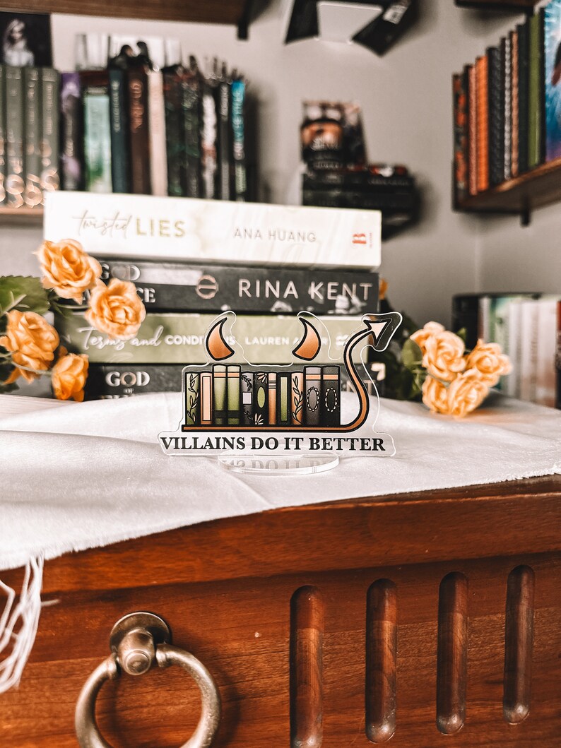 Puede incluir: Un separador de libros de acr&iacute;lico transparente con una pila de libros y una cola de diablo. El separador de libros tiene el texto "VILLAINS DO IT BETTER" en la parte inferior. El separador de libros est&aacute; sobre una mesa de madera con un pa&ntilde;o blanco encima.