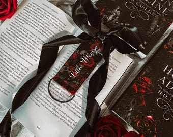 May include: A black ribbon bookmark with the words "Little Mouse" printed on it, placed on top of an open book with a red rose on the cover. The book is titled "Haunting Adeline" by H.D. Carlton.