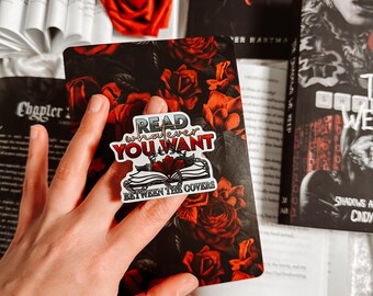 May include: A collection of books and decorative items. A sticker with the text "Read Whatever You Want Between The Covers" is placed on a book cover with a rose pattern. Red roses and open books are also visible.