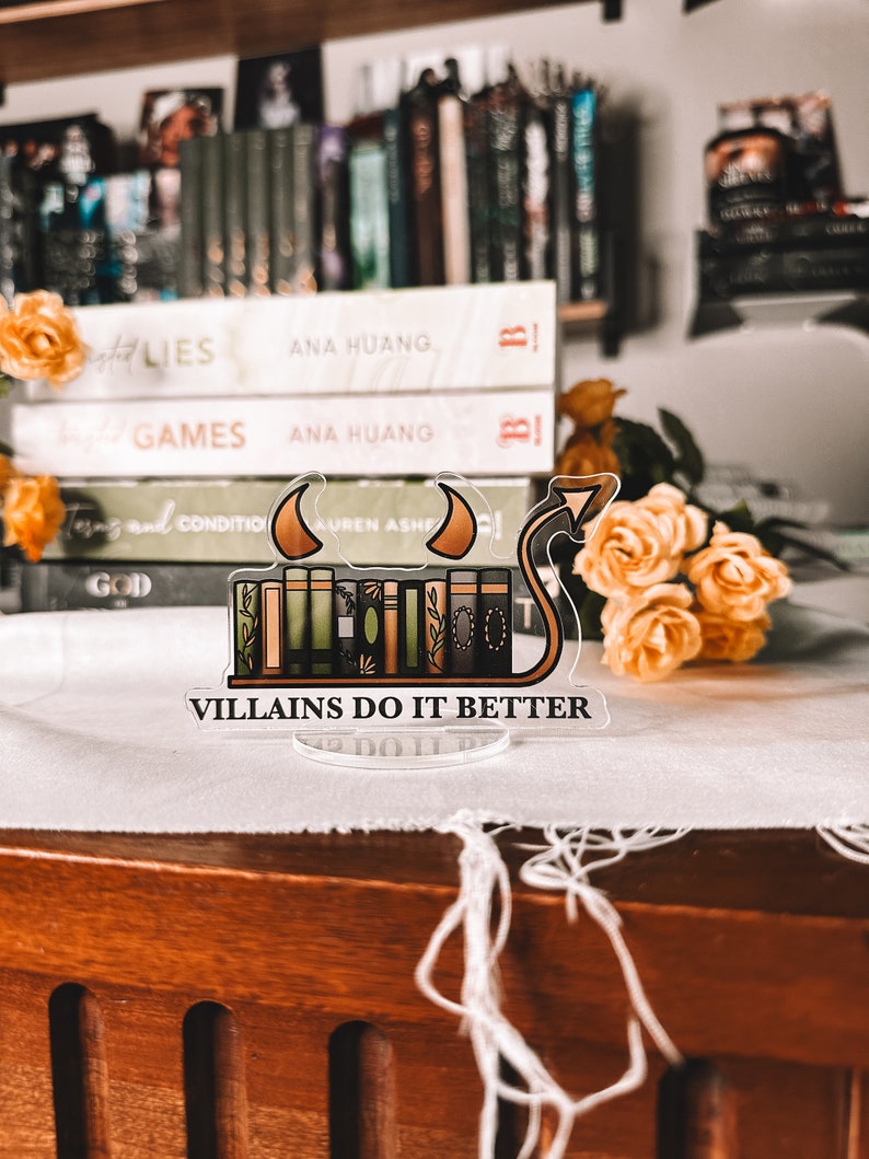 Puede incluir: Un soporte para libros de acr&iacute;lico transparente con una pila de libros y una cola de diablo. Los libros est&aacute;n apilados uno encima del otro y tienen lomos de diferentes colores. El texto "VILLAINS DO IT BETTER" est&aacute; impreso en el soporte. El soporte est&aacute; sobre una superficie blanca con algunas flores amarillas.
