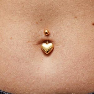 May include: A gold-colored belly button ring with a heart-shaped charm. The jewelry is shown against a background of skin and a gray shirt.