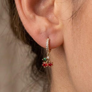 May include: A gold hoop earring featuring a small dangling cherry charm. The hoop is encrusted with tiny clear stones. The cherry charm consists of two round red stones representing the fruit and a single green stone for the leaf.