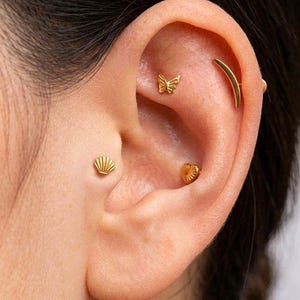 May include: Close-up of an ear adorned with multiple gold earrings. The earrings include a butterfly, a shell, a heart, and a curved bar. The jewelry is small and delicate, adding a touch of elegance.