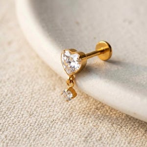 Puede incluir: Un pendiente de botón con parte posterior plana en tono dorado, que presenta una piedra preciosa transparente en forma de corazón en la parte superior y una piedra redonda más pequeña que cuelga de un delicado eslabón dorado.