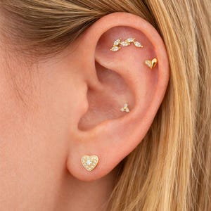 Pode incluir: Close-up de uma orelha adornada com vários brincos de ouro. Um brinco de botão em forma de coração está no lóbulo e um brinco de botão em forma de coração está na parte superior da orelha. Um grupo de três pequenos pregos e uma barra curva de pedras em forma de marquesa completam as joias de orelha.