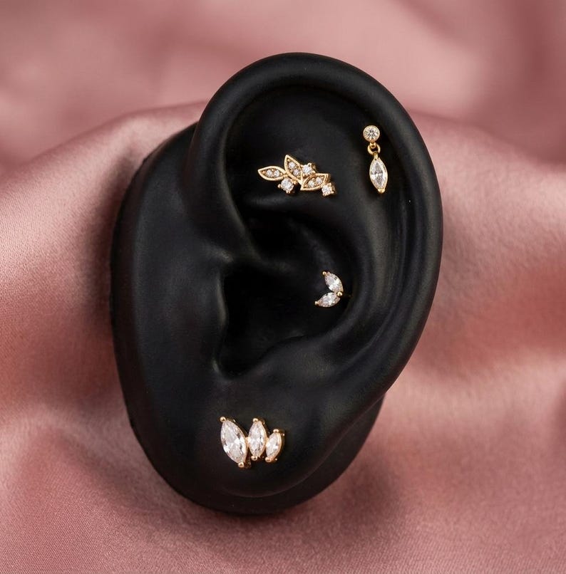 Puede incluir: Una exhibici&oacute;n de piercings dorados con piedras claras y brillantes. Los piercings incluyen una l&aacute;grima colgante, una media luna y un grupo de piedras con corte marquesa. La oreja es negra, sobre un fondo rosa.