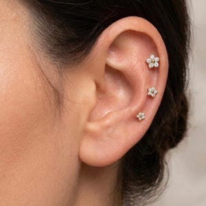 May include: Three gold-toned flower-shaped stud earrings are arranged vertically along the cartilage of an ear. Each earring features a central gold-toned bead surrounded by six small, clear, sparkling stones arranged in a floral pattern.