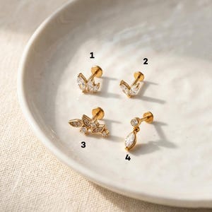 Puede incluir: Cuatro pendientes dorados con gemas claras y brillantes. Los pendientes presentan varios dise&ntilde;os, incluyendo formas de hojas y una l&aacute;grima colgante. Las joyas se exhiben sobre una superficie blanca ligeramente curvada.