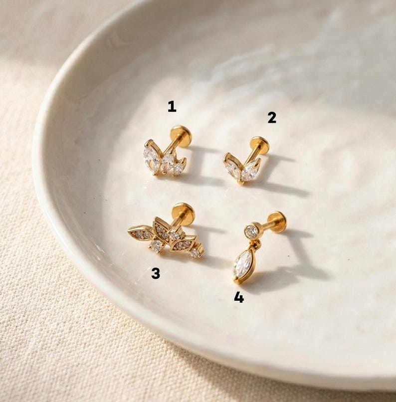 Puede incluir: Cuatro pendientes dorados con gemas claras y brillantes. Los pendientes presentan varios dise&ntilde;os, incluyendo formas de hojas y una l&aacute;grima colgante. Las joyas se exhiben sobre una superficie blanca ligeramente curvada.