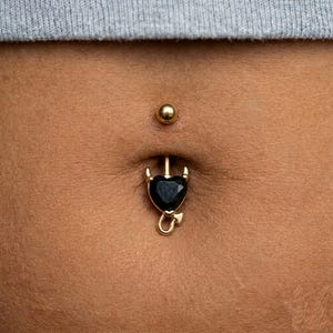 Può includere: Un piercing all'ombelico color oro con una gemma nera a forma di cuore e un ciondolo a forma di coda di diavolo. Anche il perno superiore è dorato. Il gioiello è posto su uno sfondo color carne.